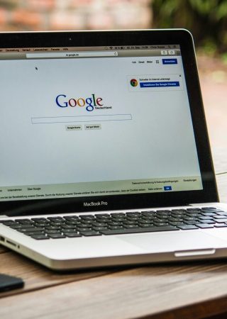 A MacBook Pro displaying Google Search on a wooden table outdoors, next to a smartphone.